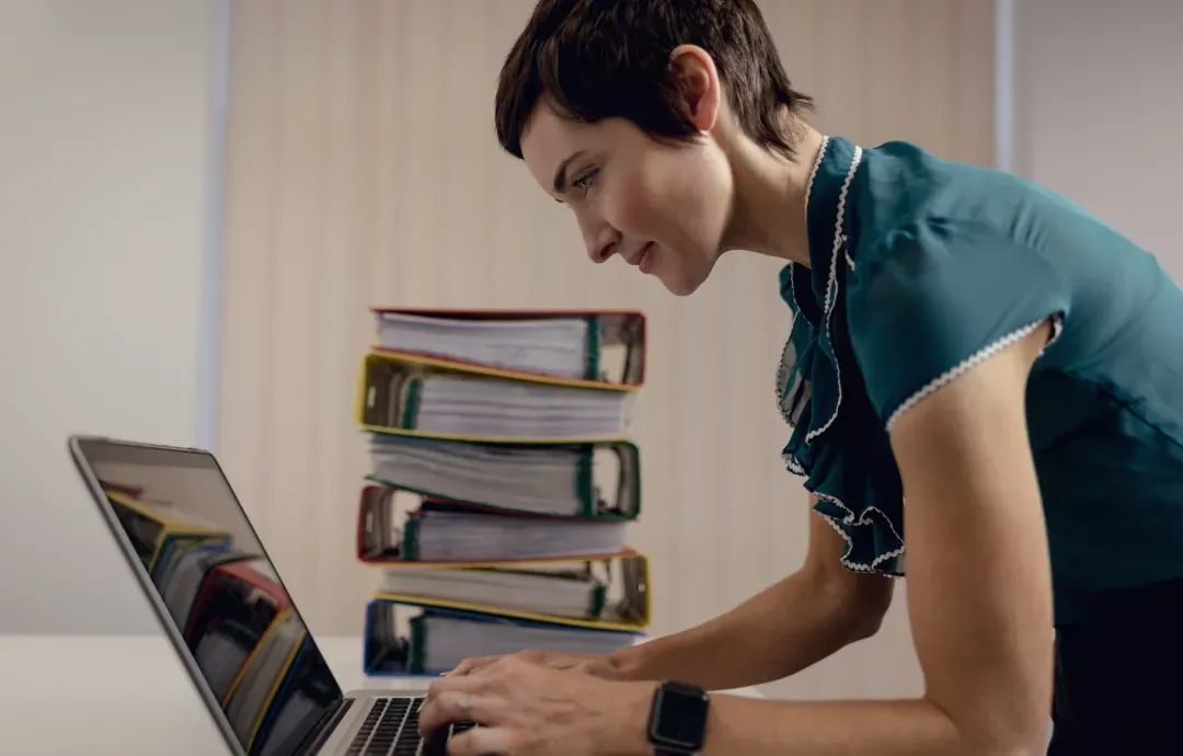A woman typing on a laptop next to a tall stack of colorful binders in an office