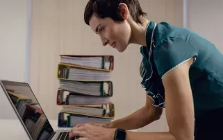 Eine Frau tippt auf einem Laptop neben einem großen Stapel bunter Aktenordner in einem Büro