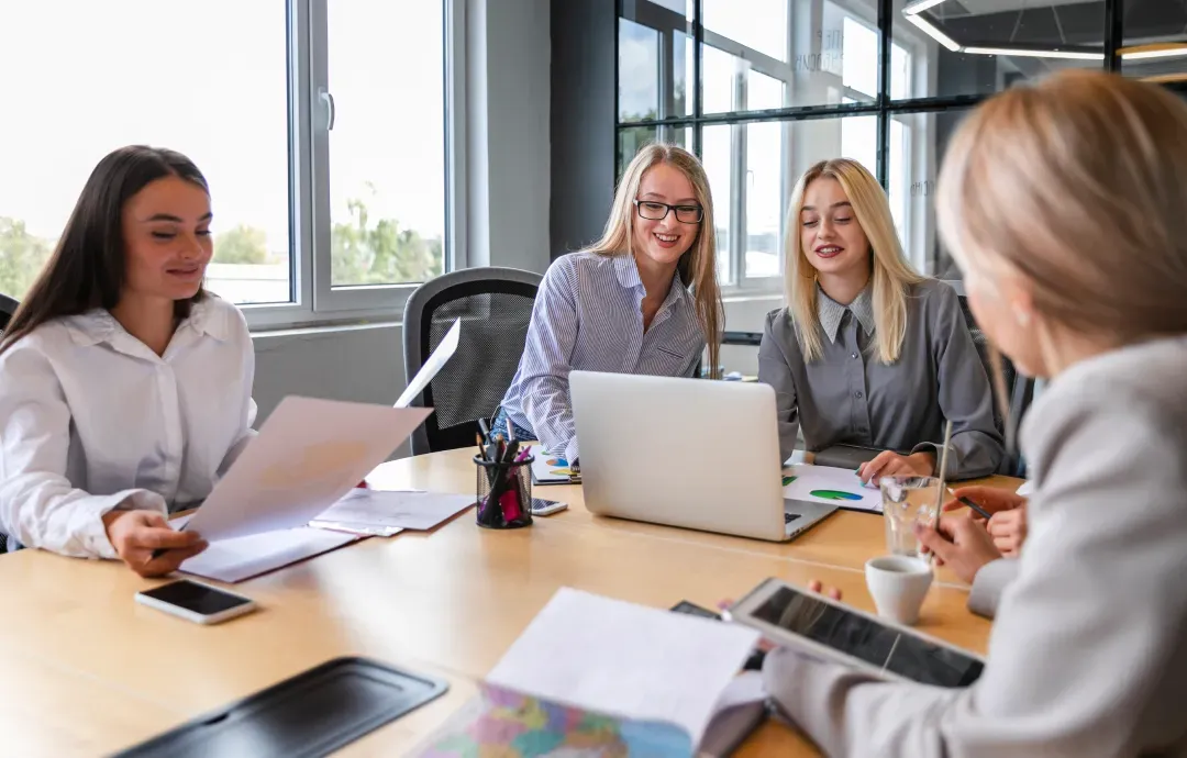 Four female colleagues smiling and reviewing documents together on a laptop during a team meeting