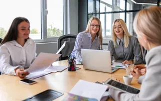 Four female colleagues smiling and reviewing documents together on a laptop during a team meeting