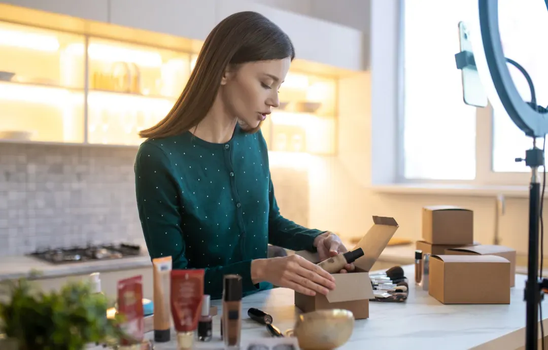 A woman packing beauty products into small cardboard boxes for online orders while filming a video with a ring light