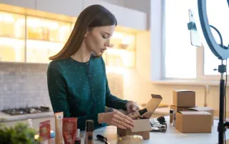 A woman packing beauty products into small cardboard boxes for online orders while filming a video with a ring light